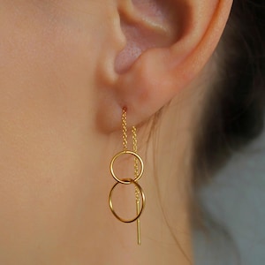 Könnte beinhalten: Nahaufnahme eines goldfarbenen Ohrrings mit zwei verbundenen Kreishalbreifen und einer Kette. Der Ohrring wird im Ohr getragen und hängt herunter. Der Ohrring besteht aus einem glänzenden, reflektierenden Metall.