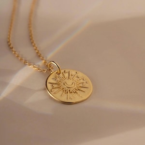 May include: A gold-toned necklace featuring a circular pendant with an embossed design of a central crescent moon surrounded by radiating sun rays and small stars. The pendant hangs from a delicate gold-toned chain with small, uniform links.