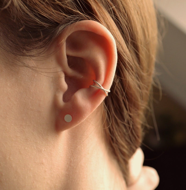 May include: A silver ear cuff and a small silver stud earring are shown on an ear. The ear cuff has a crisscross design. The earring is a simple, round stud. The skin tone is fair, and the hair is brown.