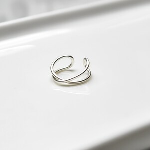May include: A silver-colored, open-ended ring with a double-band design. The ring features a smooth, polished finish and a minimalist aesthetic. The ring is designed to be worn on a finger.
