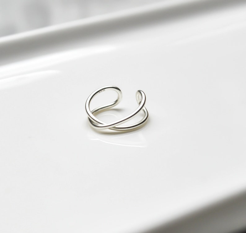 May include: A silver-colored, open-ended ring with a double-band design. The ring features a smooth, polished finish and a minimalist aesthetic. The ring is designed to be worn on a finger.