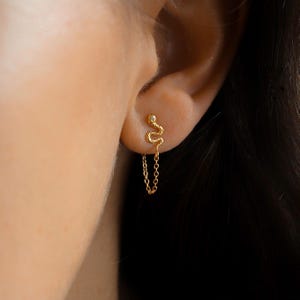 Könnte beinhalten: Ein goldfarbener Ohrring in Schlangenform mit einem kleinen Edelstein am Kopf. Der Ohrring hängt mit einer zarten Kette am Ohr. Der Ohrring wird am Ohr einer Person getragen.