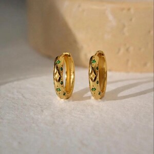 May include: Two gold-colored hoop earrings with green and diamond-shaped accents. The earrings are on a white surface. The earrings are a jewelry item.