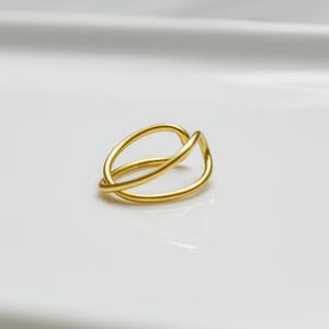 May include: A gold ring with an unusual, interwoven design. The ring has two overlapping, curved bands forming an open, abstract shape. The ring is set against a plain white background, emphasising its simple elegance and modern style.