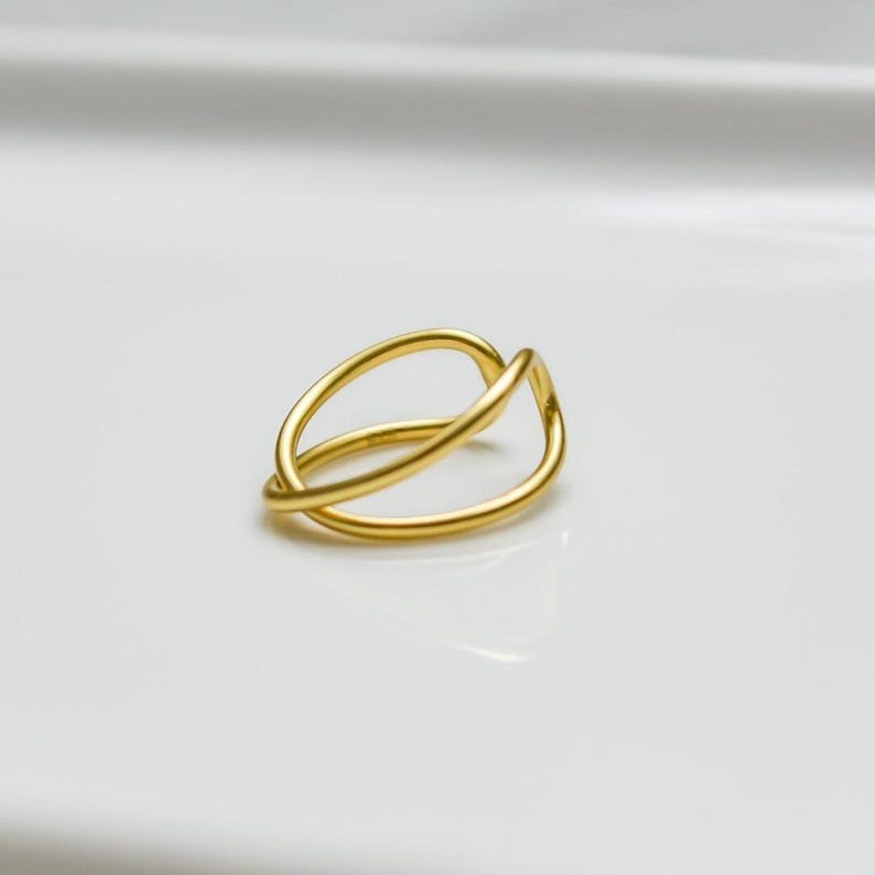May include: A gold ring with an unusual, interwoven design. The ring has two overlapping, curved bands forming an open, abstract shape. The ring is set against a plain white background, emphasising its simple elegance and modern style.