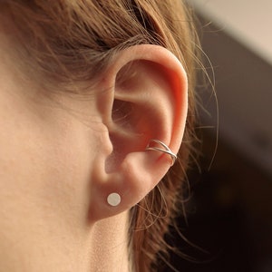 May include: A close-up of an ear with a silver ear cuff and a small, round stud earring. The ear cuff wraps around the upper ear, and the stud is in the earlobe. The skin is fair-toned, and the hair is light brown.