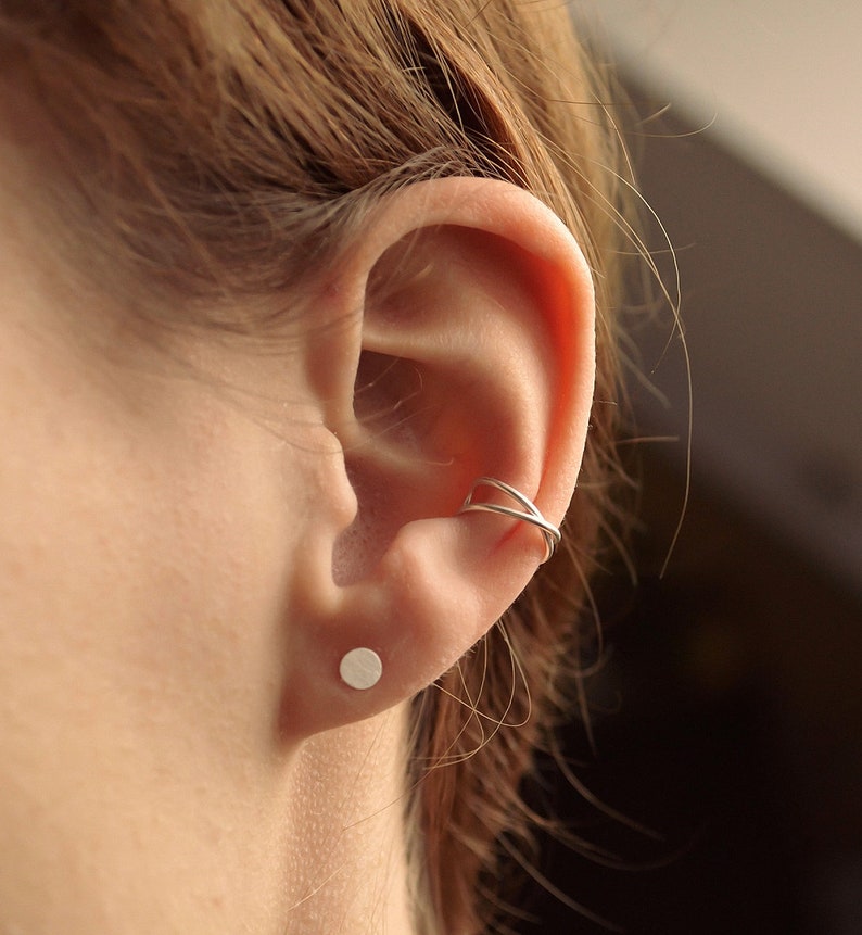 May include: A close-up of an ear with a silver ear cuff and a small, round stud earring. The ear cuff wraps around the upper ear, and the stud is in the earlobe. The skin is fair-toned, and the hair is light brown.