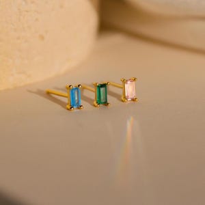 Könnte beinhalten: Drei goldene Bar-Ohrringe mit verschiedenen Edelsteinen. Die Ohrringe sind von links nach rechts angeordnet: hellblau, grün und rosa.