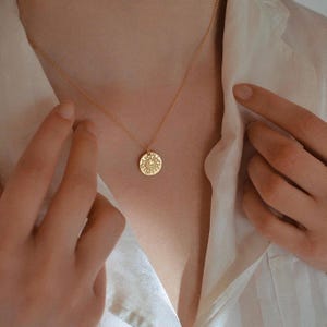 May include: A gold-toned necklace featuring a delicate chain and a small, circular pendant. The pendant displays an engraved sunburst design with a crescent moon at its center, suspended from a simple, thin metal link chain worn around the neck.