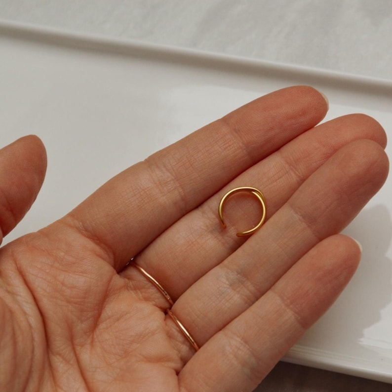 May include: A small, open gold ring. The ring is a simple, minimalist design, possibly for the ear or nose. The ring is held in a hand, with other rings visible on the fingers.