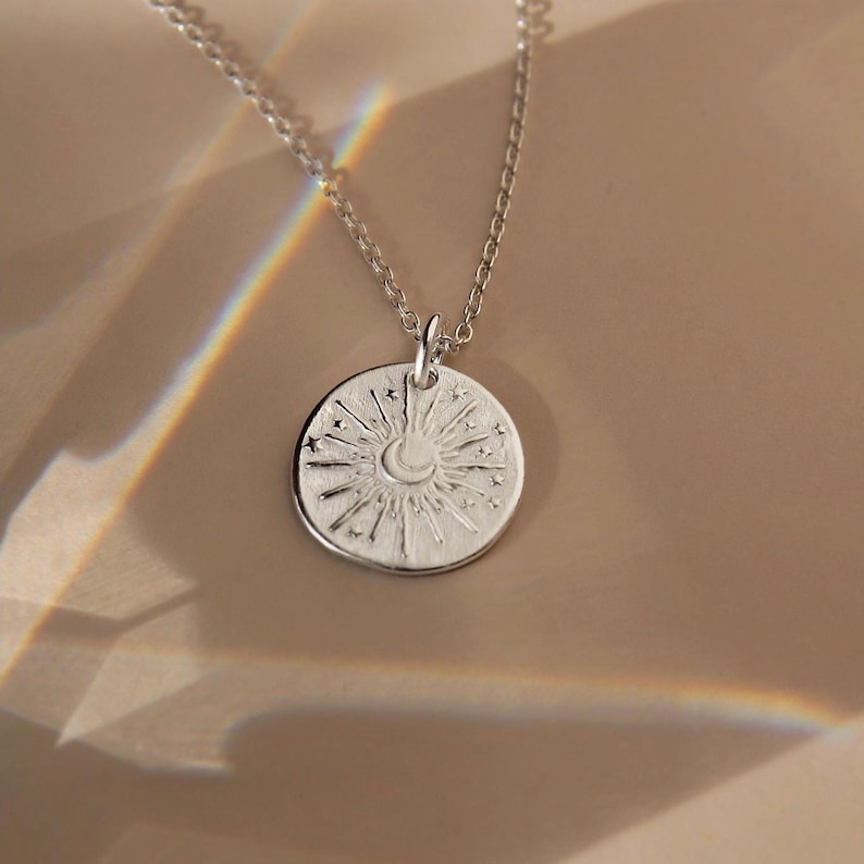 May include: A silver-toned necklace featuring a circular pendant with an embossed design of a central crescent moon surrounded by radiating sunbeams and small stars. The pendant hangs from a delicate silver-toned chain link necklace.