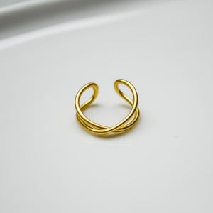 May include: A gold-coloured, open-ended ring with a crisscross design. The ring has a smooth, polished finish and is set against a white background. The ring's design features two intertwined bands that create an elegant, minimalist look.
