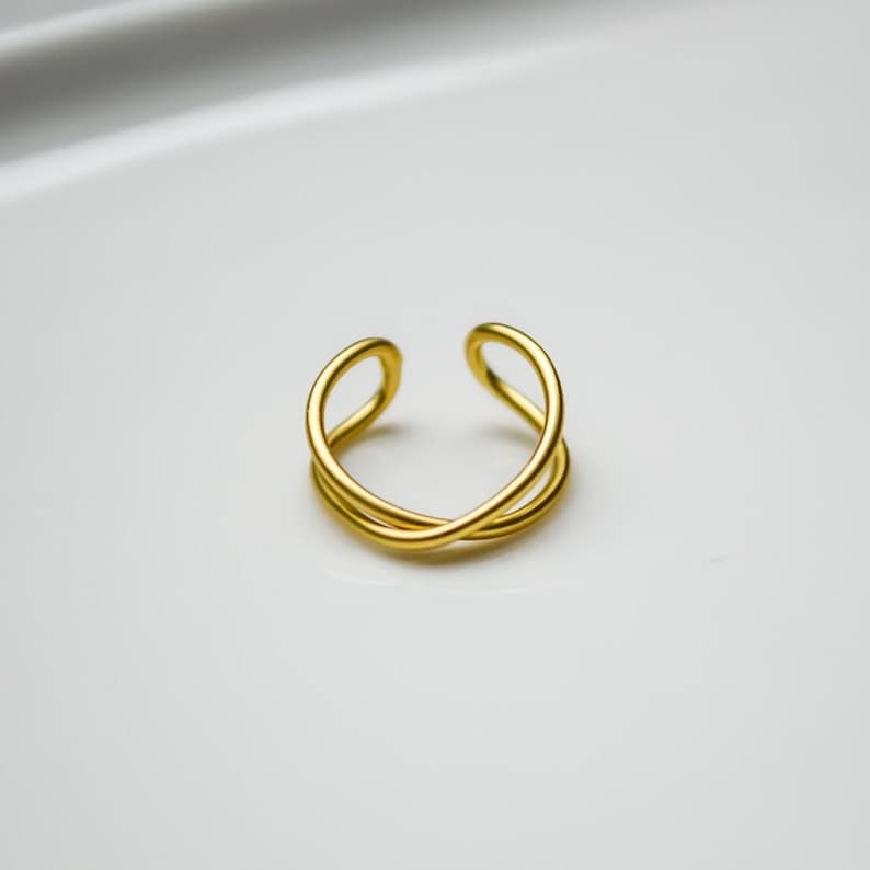 May include: A gold-coloured, open-ended ring with a crisscross design. The ring has a smooth, polished finish and is set against a white background. The ring's design features two intertwined bands that create an elegant, minimalist look.