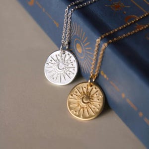 May include: Two circular pendant necklaces, one in silver and one in gold, each featuring an embossed sunburst design with a central crescent moon and small stars. The pendants hang from delicate matching metal chains, showcasing a detailed celestial motif.