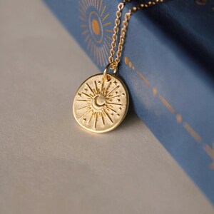 May include: A gold-toned necklace featuring a circular pendant with an embossed sun and crescent moon design surrounded by small stars. The pendant hangs from a delicate gold-toned chain, showcasing a textured finish on the metal surface.