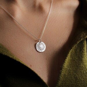 May include: A silver necklace featuring a small, round pendant with an engraved sun and moon design. The pendant is suspended from a delicate silver chain, showcasing intricate rays and star motifs etched into the circular metal surface.