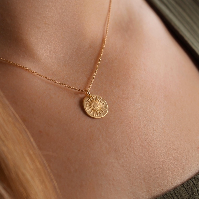 May include: A gold-toned necklace featuring a delicate chain and a circular pendant. The pendant displays an embossed design of a central crescent moon surrounded by radiating sunbeams and small dots, creating a celestial-inspired motif.