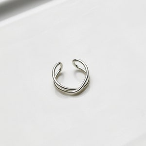 May include: A silver-colored, open-ended ring with a minimalist design. The ring features a continuous, intertwined loop, creating an elegant and modern aesthetic. The ring is shown on a white surface.