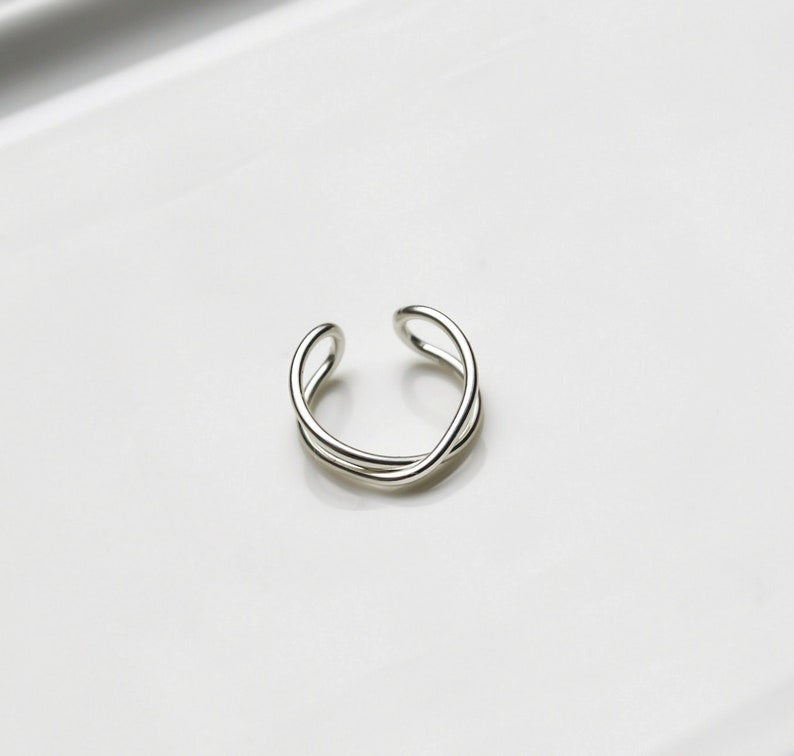 May include: A silver-colored, open-ended ring with a minimalist design. The ring features a continuous, intertwined loop, creating an elegant and modern aesthetic. The ring is shown on a white surface.