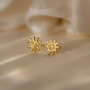 Könnte beinhalten: Ein Paar goldfarbene Ohrstecker. Jeder Ohrring hat ein Sonnenmuster mit einer zentralen Spirale. Die Ohrringe sind vor einem weichen, neutralen Hintergrund platziert, der ihre helle, metallische Oberfläche hervorhebt.