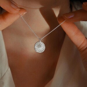 May include: A silver-toned necklace featuring a circular pendant with an embossed design of a crescent moon surrounded by radiating sunbeams and small star-like dots. The pendant hangs from a delicate, thin silver-toned metal chain link necklace.