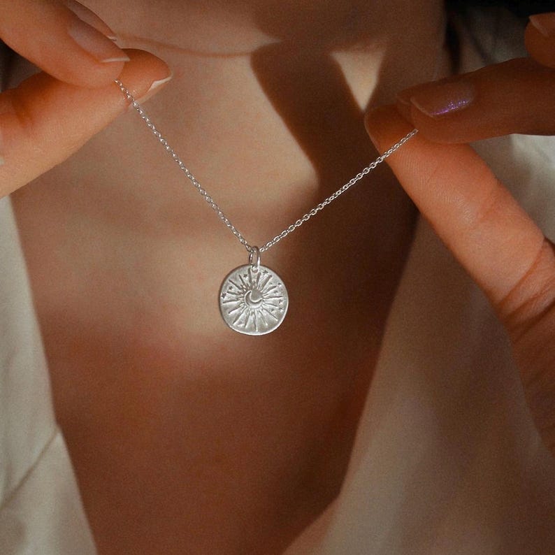 May include: A silver-toned necklace featuring a circular pendant with an embossed design of a crescent moon surrounded by radiating sunbeams and small star-like dots. The pendant hangs from a delicate, thin silver-toned metal chain link necklace.