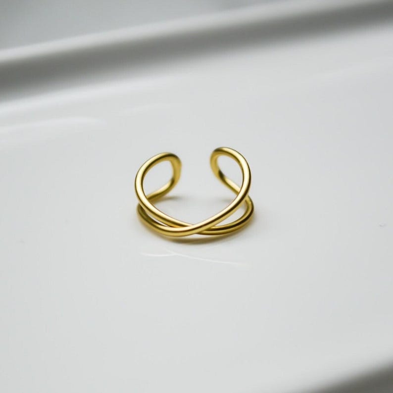 May include: A gold-coloured, open-ended ring with a double-band design. The ring features a crisscross pattern, creating an elegant and minimalist aesthetic. The jewellery piece is set against a white background.