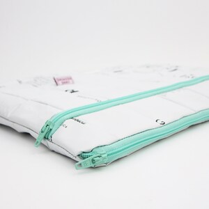 Könnte beinhalten: Eine weiße Reißverschlusstasche mit einem mintgrünen Reißverschluss. Die Tasche ist aus recyceltem Stoff gefertigt und hat ein Muster aus Nähanweisungen.