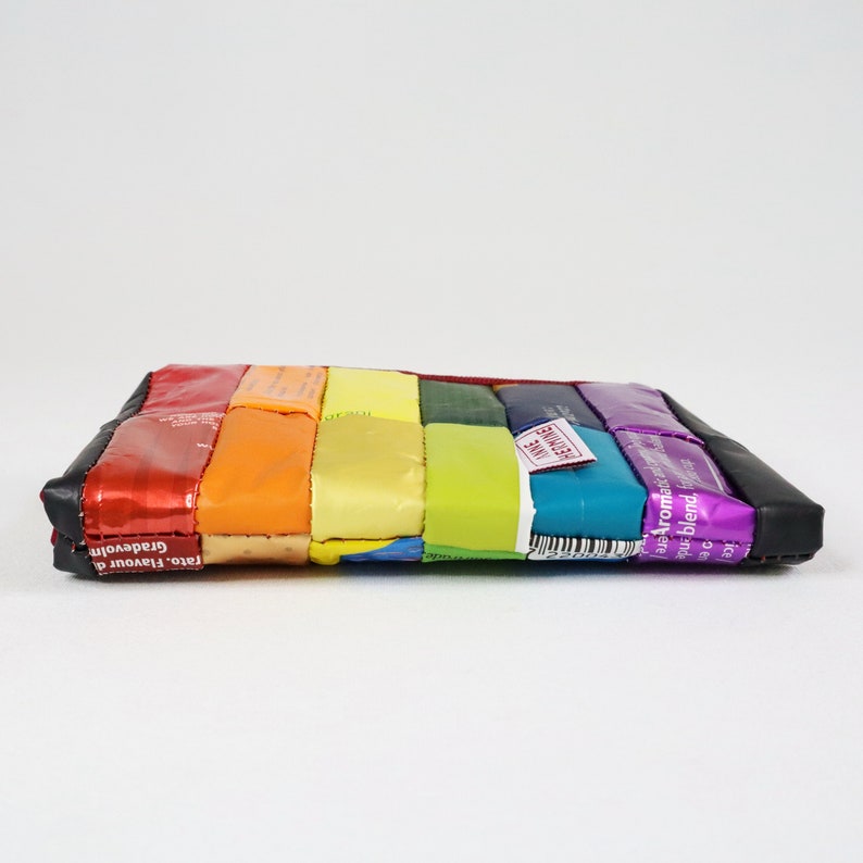 May include: A rainbow-coloured pouch made from recycled sweet wrappers. The pouch is made of different coloured wrappers, including red, orange, yellow, green, blue, and purple. The pouch has a black leather flap on the top.