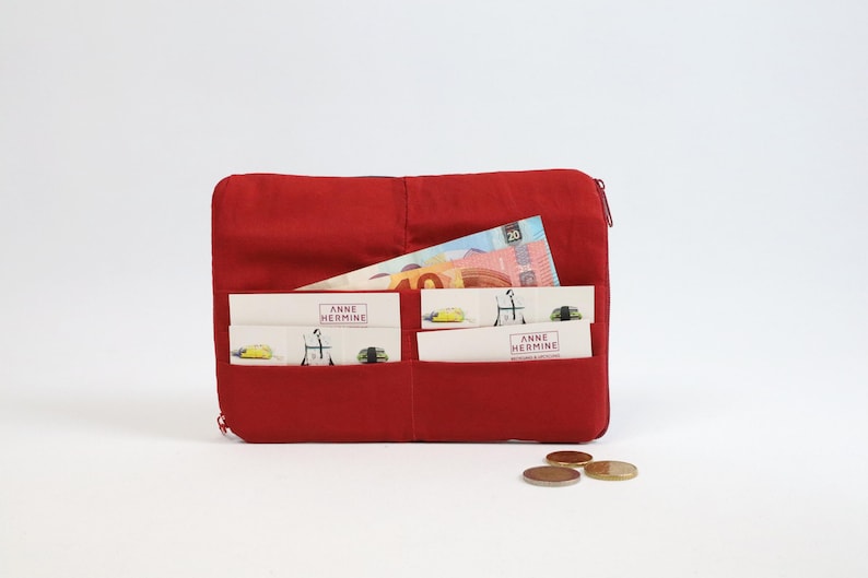May include: A red fabric wallet with multiple pockets and a zip closure. The wallet contains cash and cards with the text "Anne Hermine" printed on them.