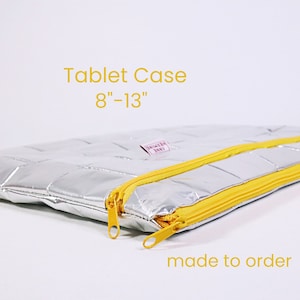 Könnte beinhalten: Silberne, gesteppte Tablettentasche mit gelben Reißverschlüssen. Die Tasche ist auf Bestellung gefertigt und passt für Tablets von 8 bis 13 Zoll.