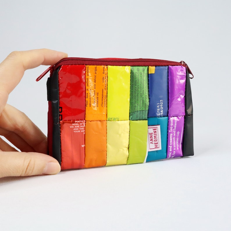 May include: A rainbow-coloured zippered pouch made from recycled packaging. The pouch is made of various colours of plastic, including red, orange, yellow, green, blue, and purple. The zipper is red. The pouch is approximately 15 cm wide and 10 cm tall.