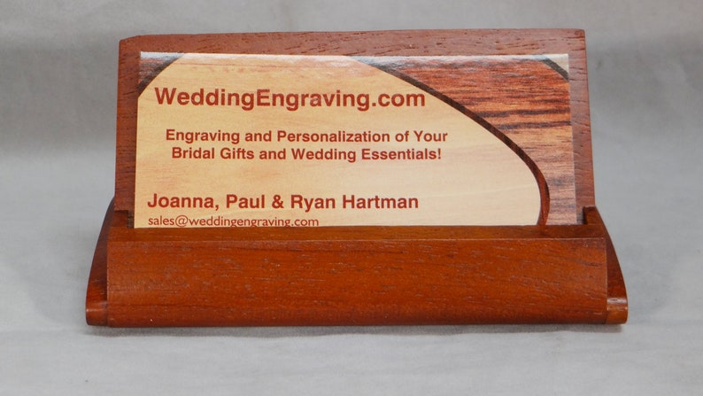 May include: A wooden business card holder with a red-brown finish. The holder has a slot for a business card and a raised section for displaying the card. The business card is for WeddingEngraving.com and features the text "Engraving and Personalization of Your Bridal Gifts and Wedding Essentials!" and "Joanna, Paul & Ryan Hartman" with the email address "sales@weddingengraving.com".