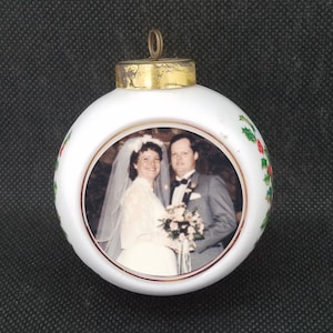 May include: White Christmas ornament with a gold top and a picture of a bride and groom in a round frame. The ornament is decorated with green holly leaves and red berries.