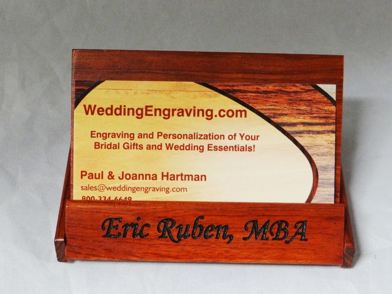 May include: A wooden business card holder with a business card inside. The business card is for WeddingEngraving.com and has the text "Engraving and Personalization of Your Bridal Gifts and Wedding Essentials!"  The business card holder has the name "Eric Ruben, MBA" engraved on the front.