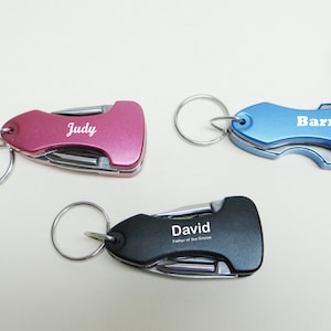 May include: Three personalized multi-tool keychains. The keychain on the left is pink with the name "Judy" engraved on it. The keychain in the middle is black with the name "David" engraved on it. The keychain on the right is blue with the name "Barry" engraved on it.