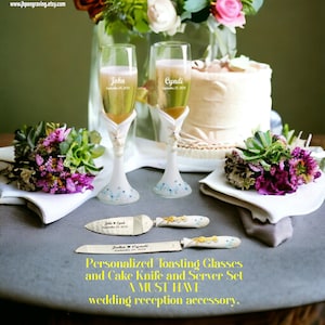 May include: Personalized toasting glasses with the names John and Cyndi engraved on them, along with a cake knife and server set with the same names. The glasses and knife are on a white table cloth with a bouquet of flowers on each side.