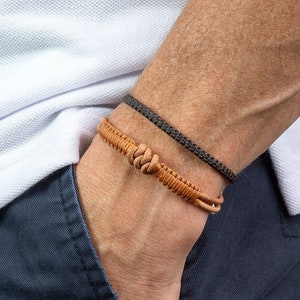 May include: Two bracelets on a wrist. One is a dark gray woven cord, and the other is a tan leather bracelet with a knotted design. The person is wearing a white polo shirt and navy blue pants.