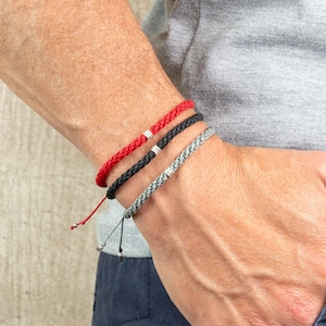 May include: Three bracelets on a person's wrist. The bracelets are red, black, and gray. The bracelets have silver accents.