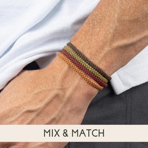 May include: A person wearing four different colored braided bracelets on their wrist. The bracelets are brown, orange, green, and dark green. The text "MIX & MATCH" is visible at the bottom of the image.