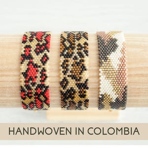 May include: Three beaded bracelets with animal print patterns in red, black, gold, and neutral tones. The bracelets are handwoven and displayed on a wooden surface. The text "HANDWOVEN IN COLOMBIA" is at the bottom.
