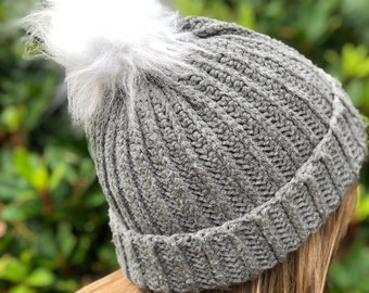 May include: A gray knit beanie with a white faux fur pom-pom. The hat is ribbed and appears to be handmade. The beanie is a winter accessory.