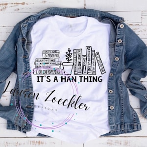 May include: White t-shirt with a black graphic design featuring stacked books, a potted plant, and the text "IT'S A HAN THING." The shirt is displayed on a denim jacket. The design includes phrases like "Fire with Fire" and "My True Love Gave to Me."