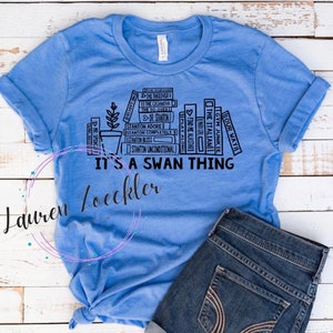 Puede incluir: Una camiseta azul claro con un gráfico de una pila de libros con títulos como "The Takeover", "The Casamova", "Dr. Stanton", "Stanton Adore", "Stanton Completely", "Stanton Bless", "Stanton Unconditional", "Hour Wait", "The Italian", "Gym Junkie", "Play Along", "Find Me Alastari" y el texto "It's a Swan Thing".