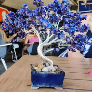 May include: A wire-wrapped tree with purple and blue gemstone leaves in a blue ceramic pot. The tree is sitting on a wooden table.