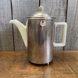 May include: A silver hammered metal teapot with a white porcelain spout and handle. The teapot has a hinged lid with a wooden knob.