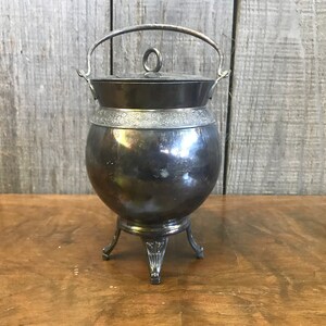 May include: A silver-plated, round, lidded container with a handle and three legs. The container has a decorative band around the middle.