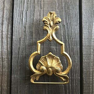 May include: Gold-toned metal towel ring with an ornate design. The ring has a scalloped shell-like center and a decorative top with a floral motif.