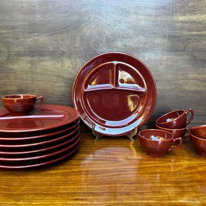 May include: A set of six red ceramic cups and a stack of six red ceramic divided plates. The plates are stacked on top of each other and the cups are arranged in a semi-circle around the plates.