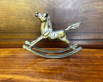 May include: A gold-colored metal rocking horse figurine. The horse is in a galloping pose with its head turned to the side. The rocking base is made of metal and has a smooth, polished finish.